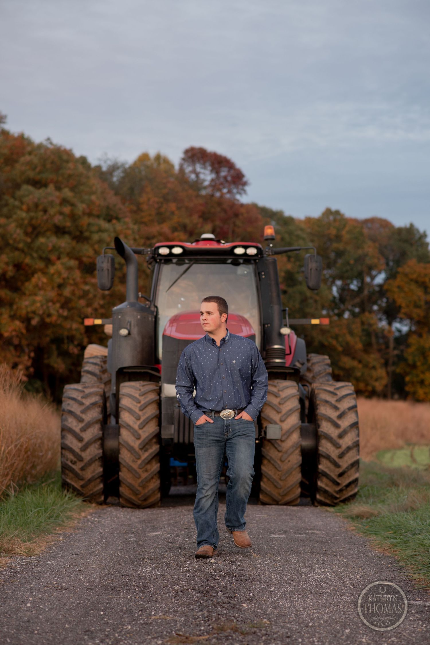 Person in casual attire poses in front of massive red tractor on rural path.