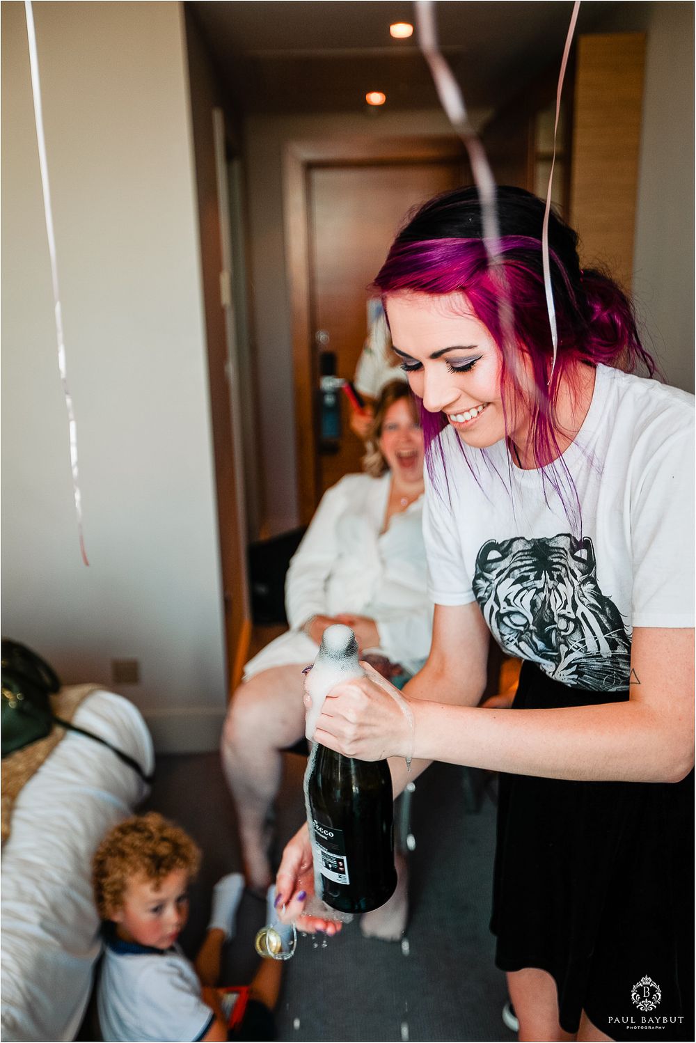 Person with bright pink hair opening champagne bottle during wedding preparations.