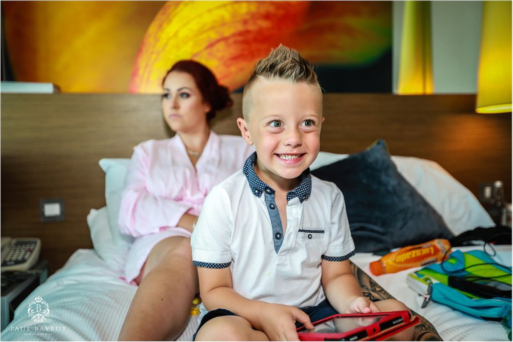 Child sitting on hotel bed with colorful backpack while someone relaxes in background.