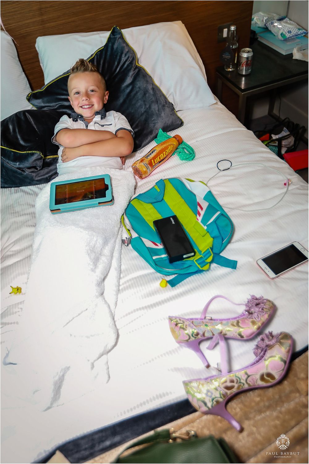 Young child relaxing on hotel bed with electronics and purple shoes visible.