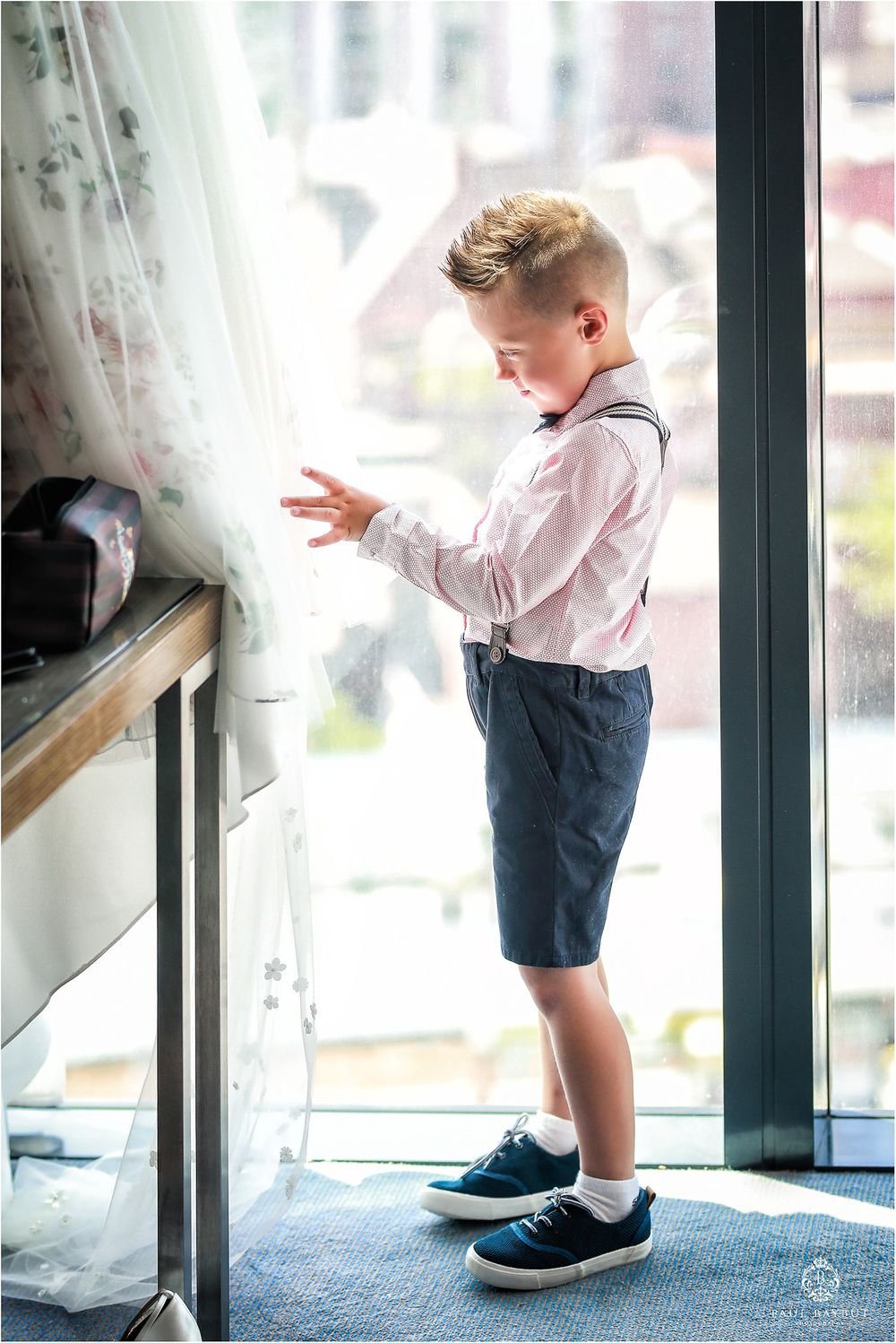 A young boy in pink dress shirt and shorts plays with balloons near a window.