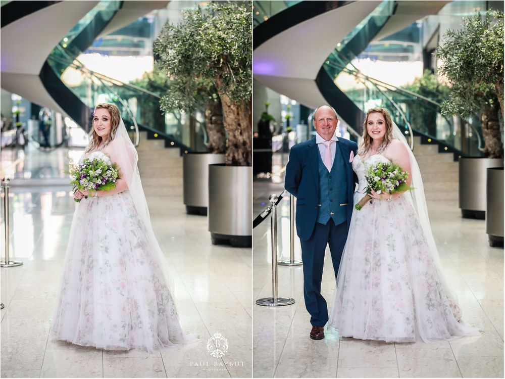 A bride in a lace wedding gown walks through a modern building hallway.