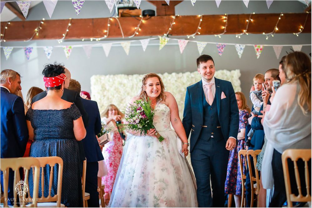 Wedding couple walks down aisle while guests in colorful attire look on.