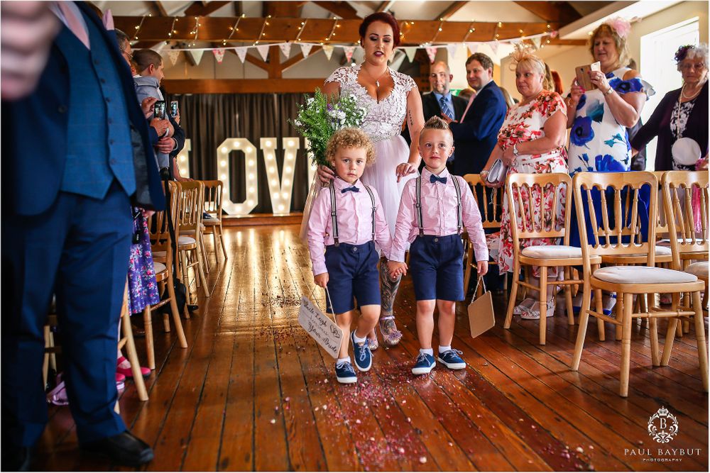 Young ring bearers in blue suspenders walk down aisle during wedding ceremony.