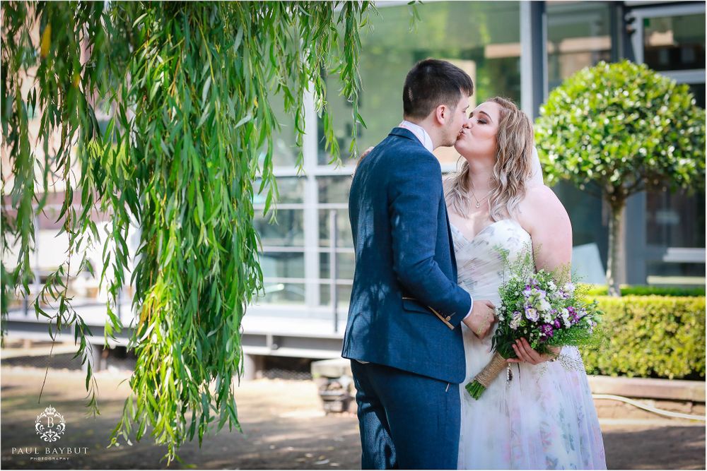 Newlyweds embrace under cascading willow branches in garden setting.