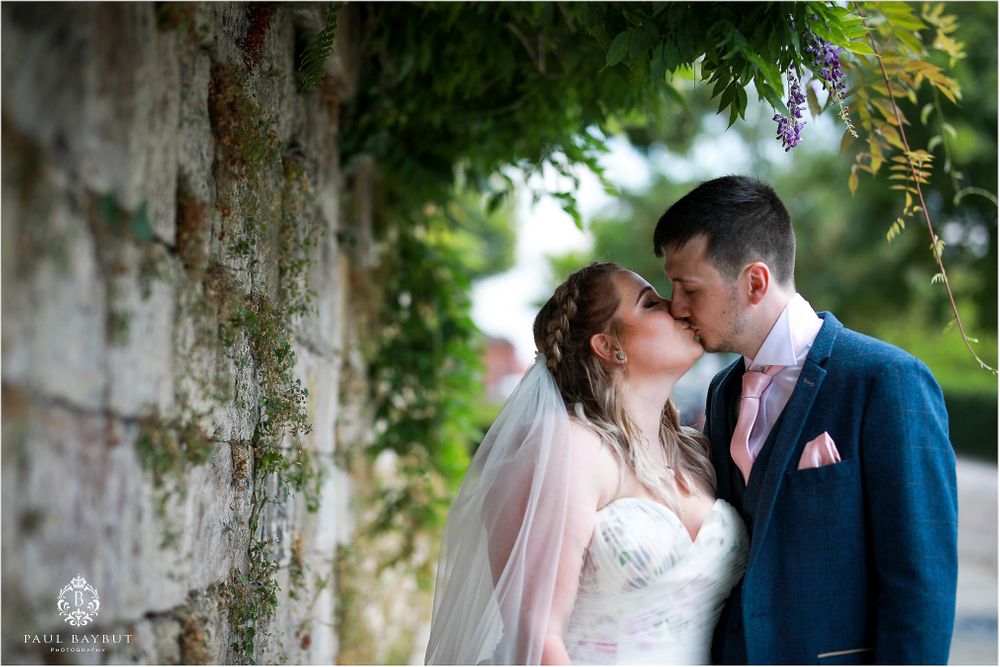 A romantic wedding moment captured under green foliage against a stone wall background.