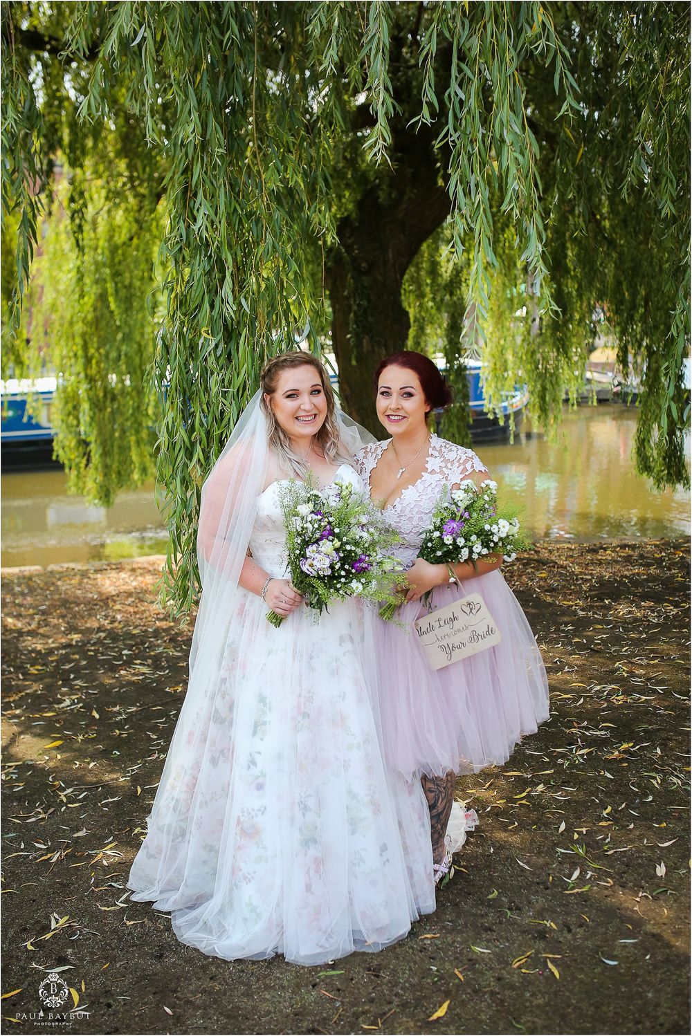 Two wedding attendees stand beneath a weeping willow tree holding colorful bouquets.