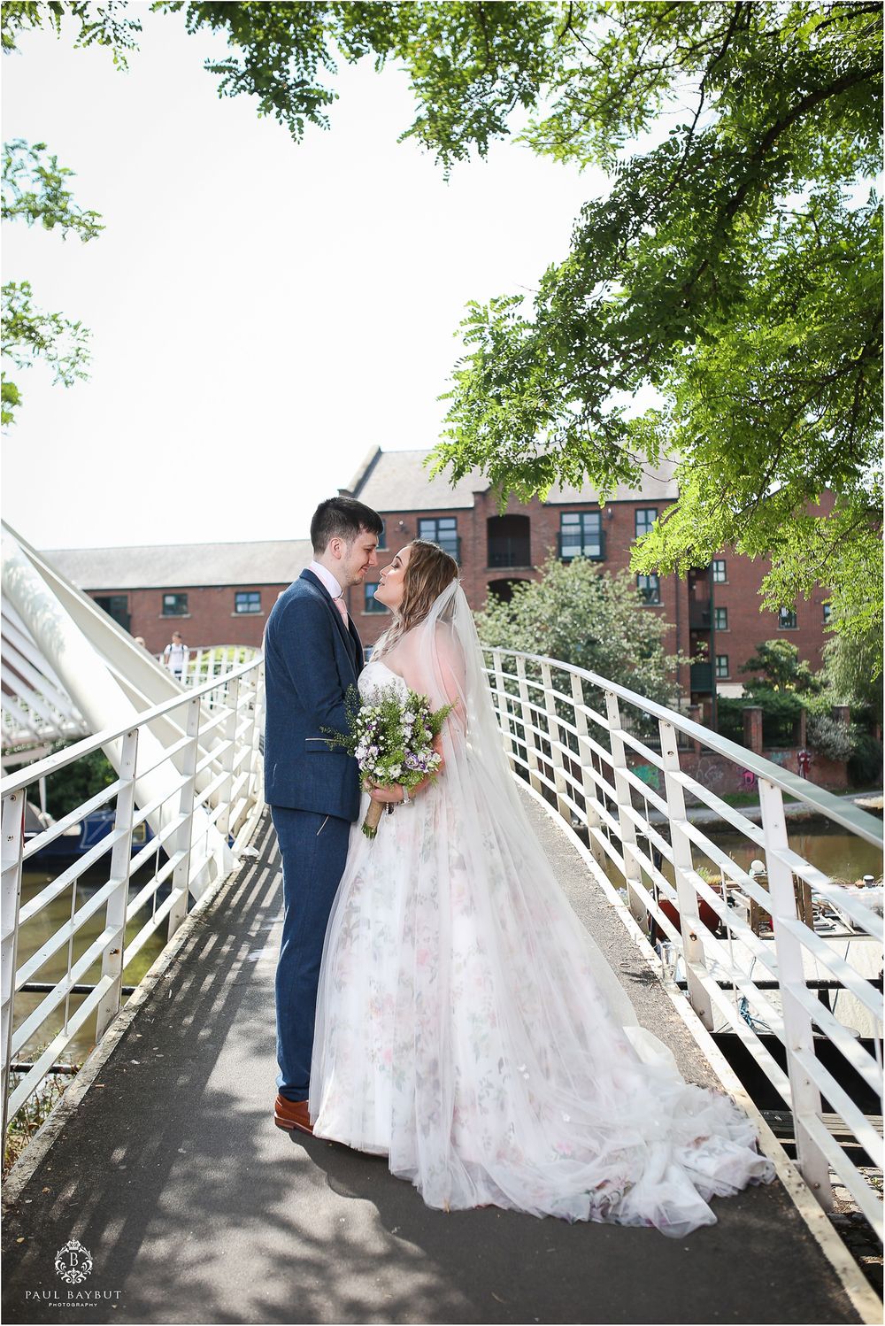 A wedding couple shares a romantic moment on a white bridge with brick buildings in background.