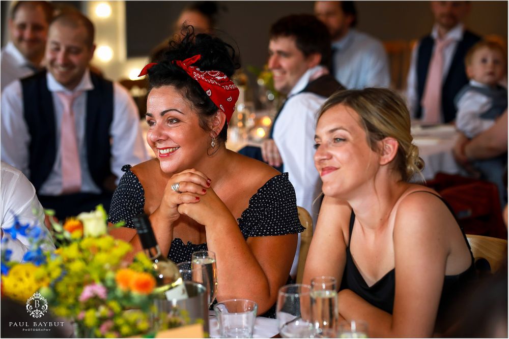 Reception guests enjoying entertainment at table decorated with bright yellow and blue flowers.