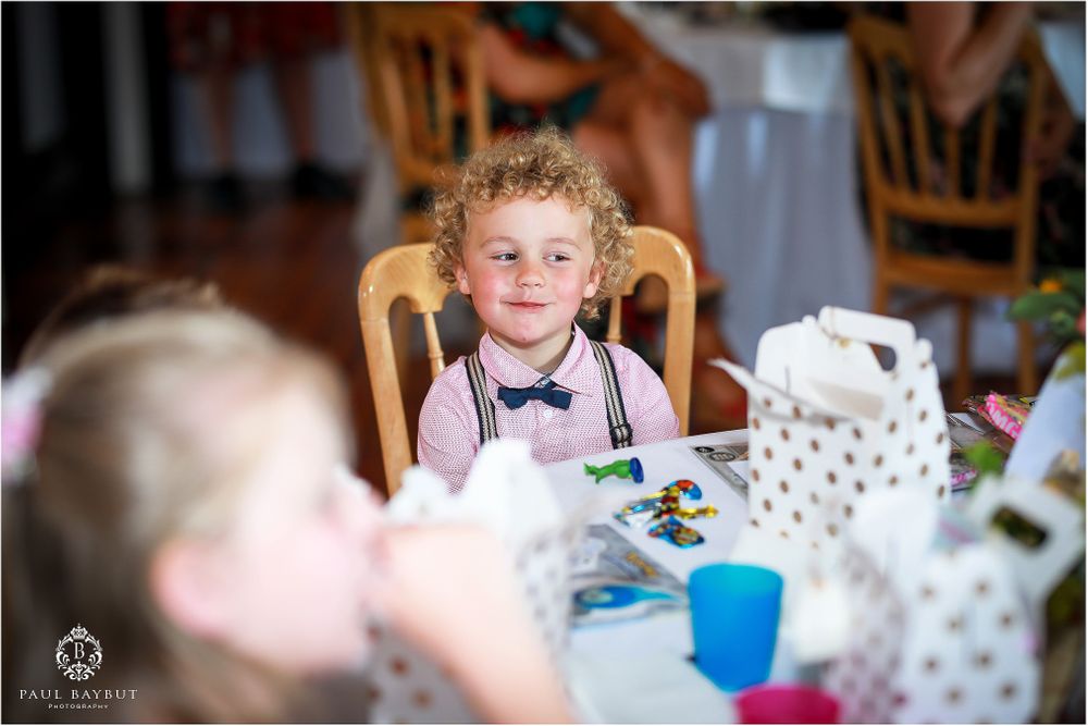 Young guest in bowtie sitting at wedding reception table with teapot and place settings.