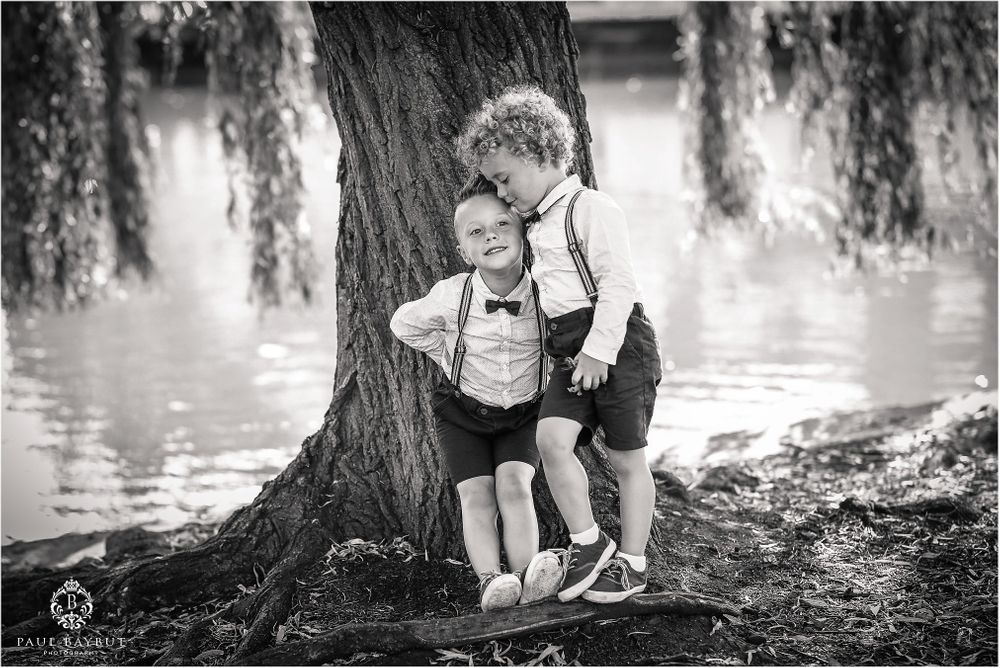 Two young children in formal attire share a tender moment by a willow tree.