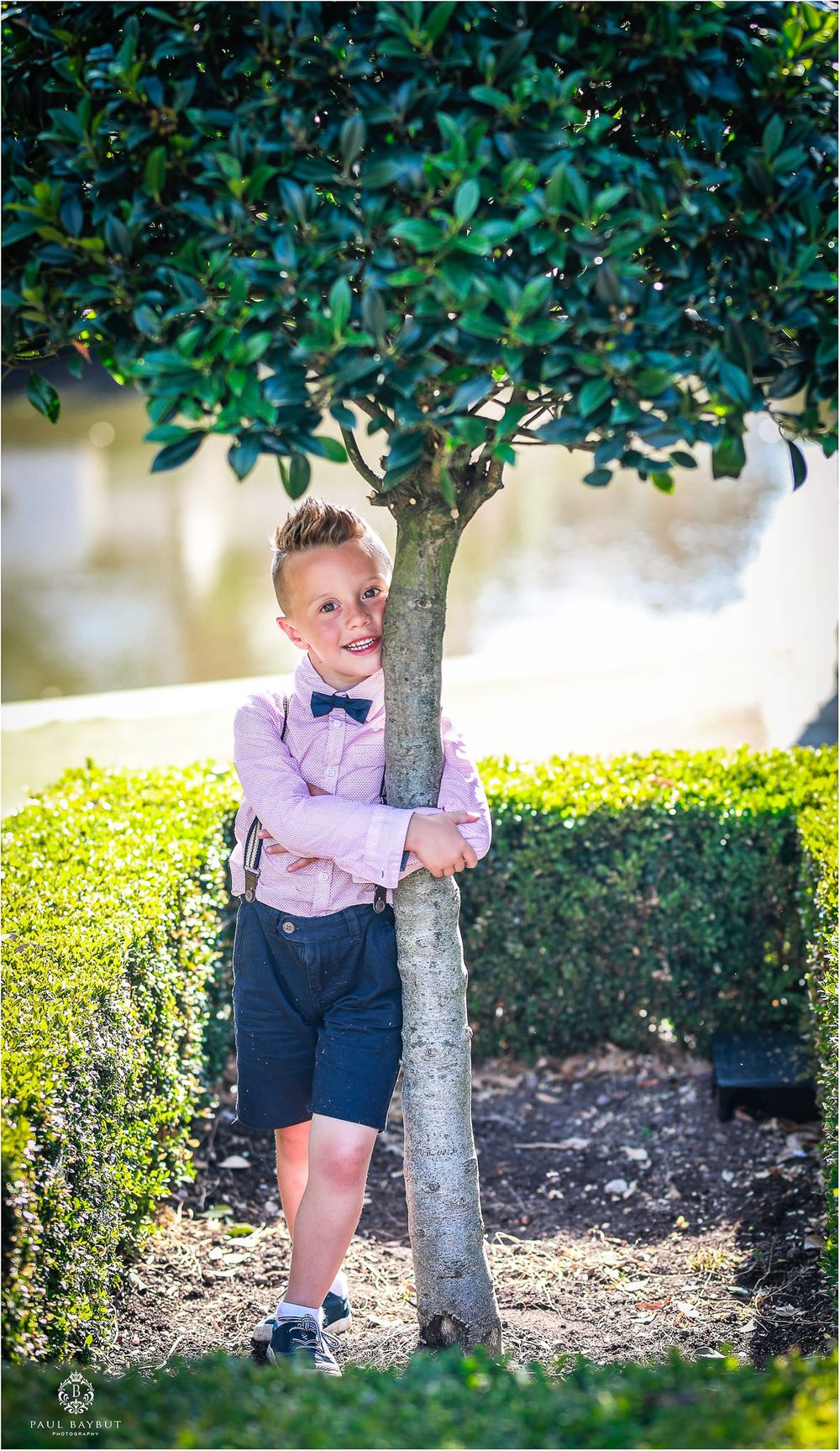 A young boy in formal attire hugs a small tree in a garden setting.
