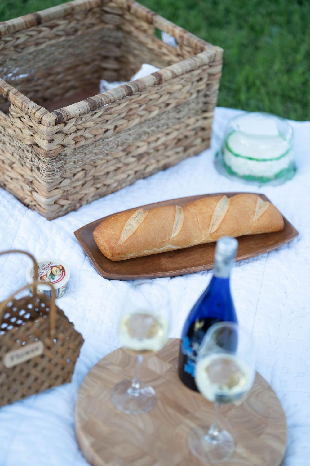 Picnic setup with wicker baskets, fresh bread, and wine glasses on white blanket.