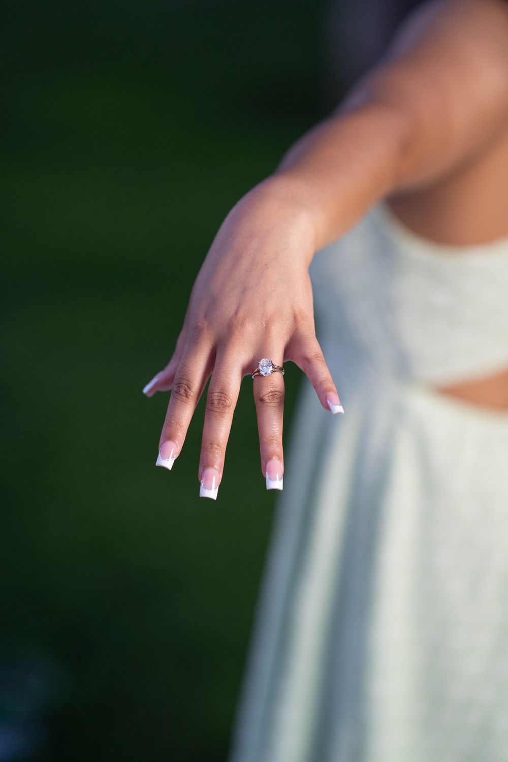 Hand showing off elegant engagement ring with manicured nails against green background.