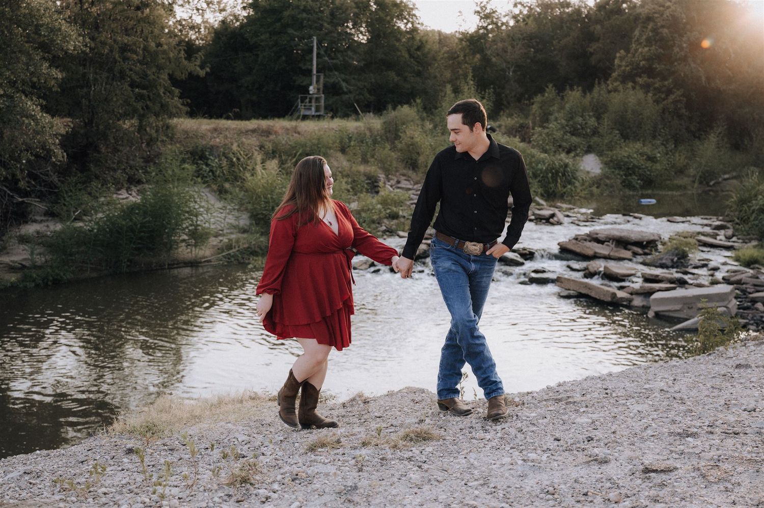 A couple walks hand in hand along a rocky riverbank at sunset.
