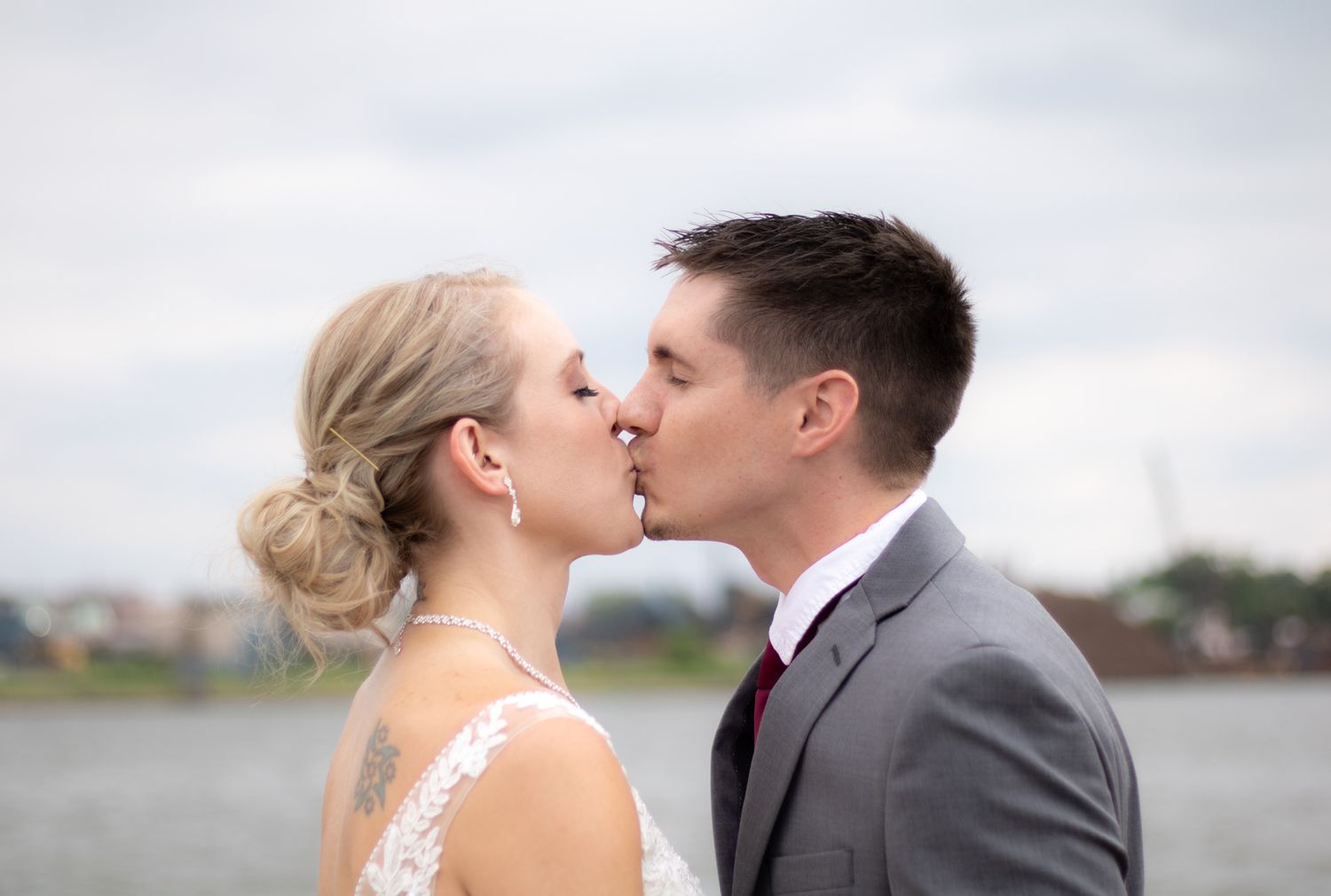 A romantic wedding photo of a couple kissing against an overcast waterfront backdrop.