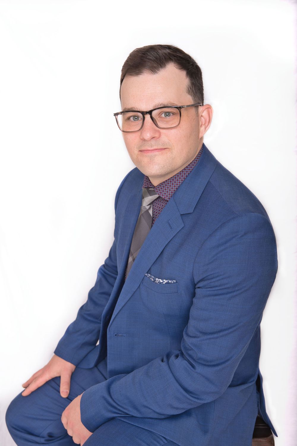Professional headshot of man in blue suit jacket and glasses against white background.