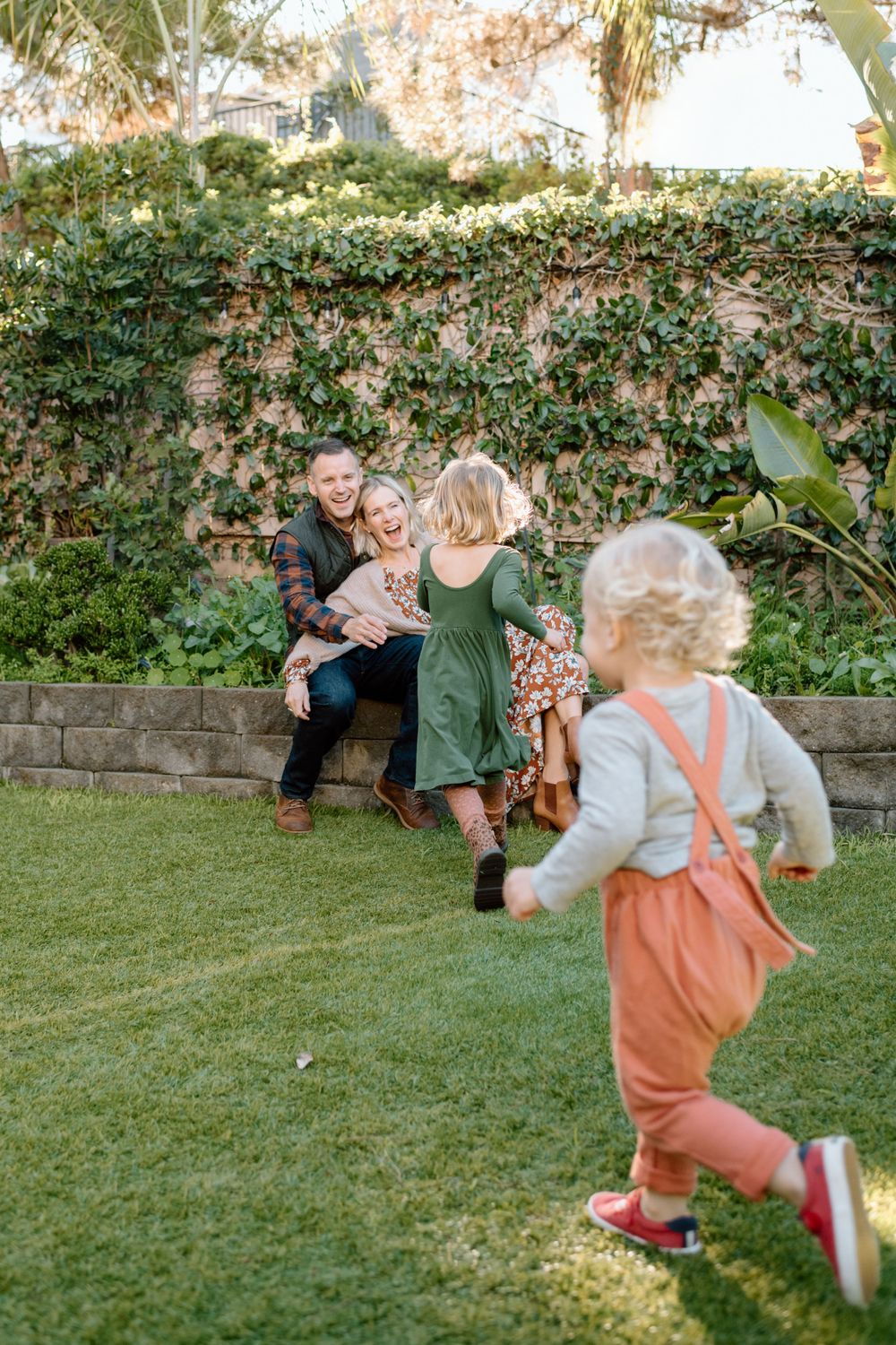 Family gathering in a garden with ivy-covered wall while a child runs on the grass during family photoshoot in San Diego.