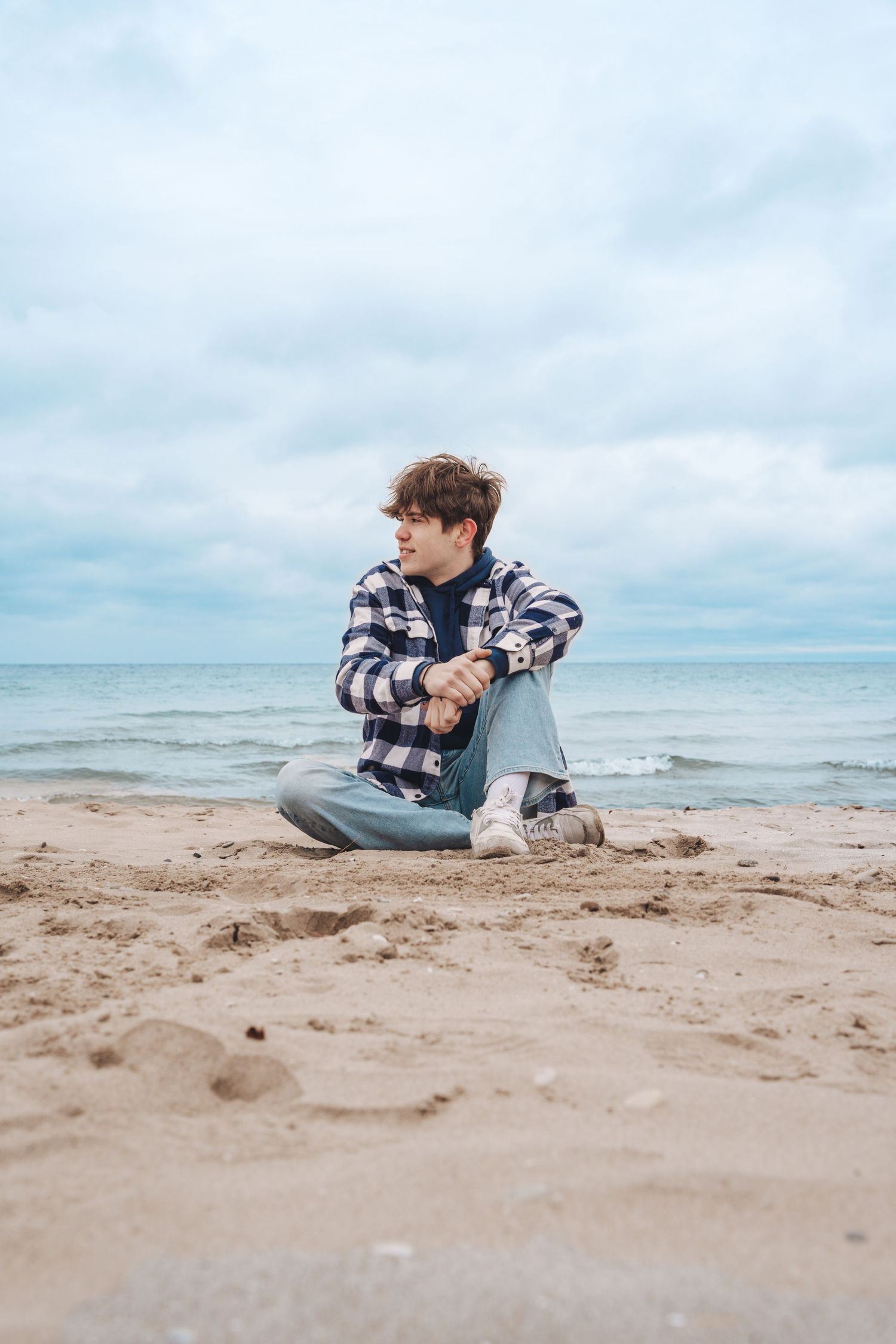 Thoughtful figure in plaid shirt sitting alone on beach sand against cloudy oceanside backdrop.
