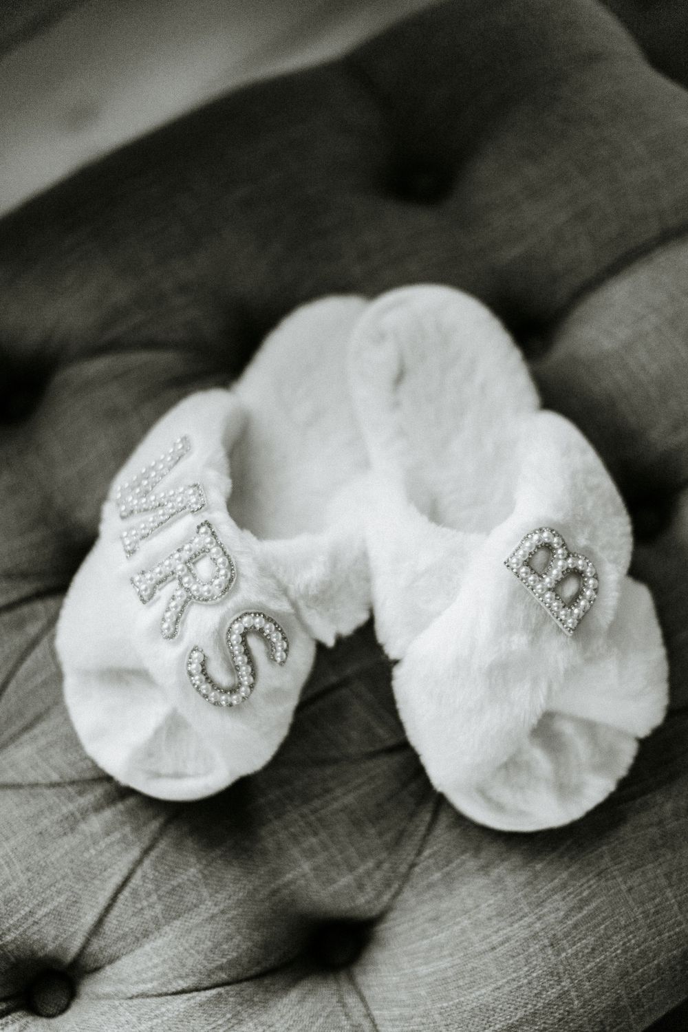 White plush slippers with crystal monogram details on grey cushioned surface.