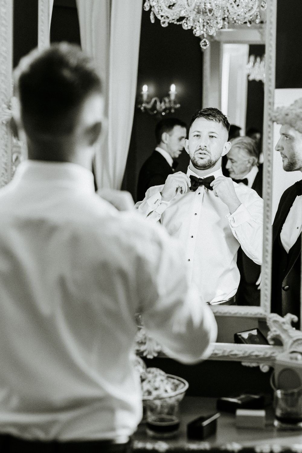 Black and white image of groomsmen helping with bow tie preparation.