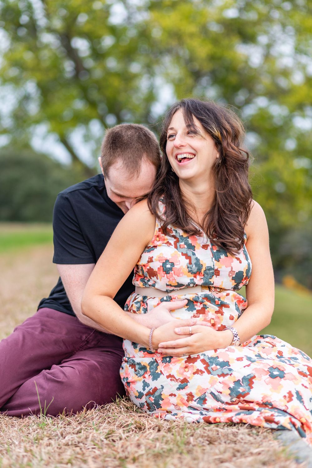 A couple shares a tender moment on grass while expecting a baby in a floral dress.