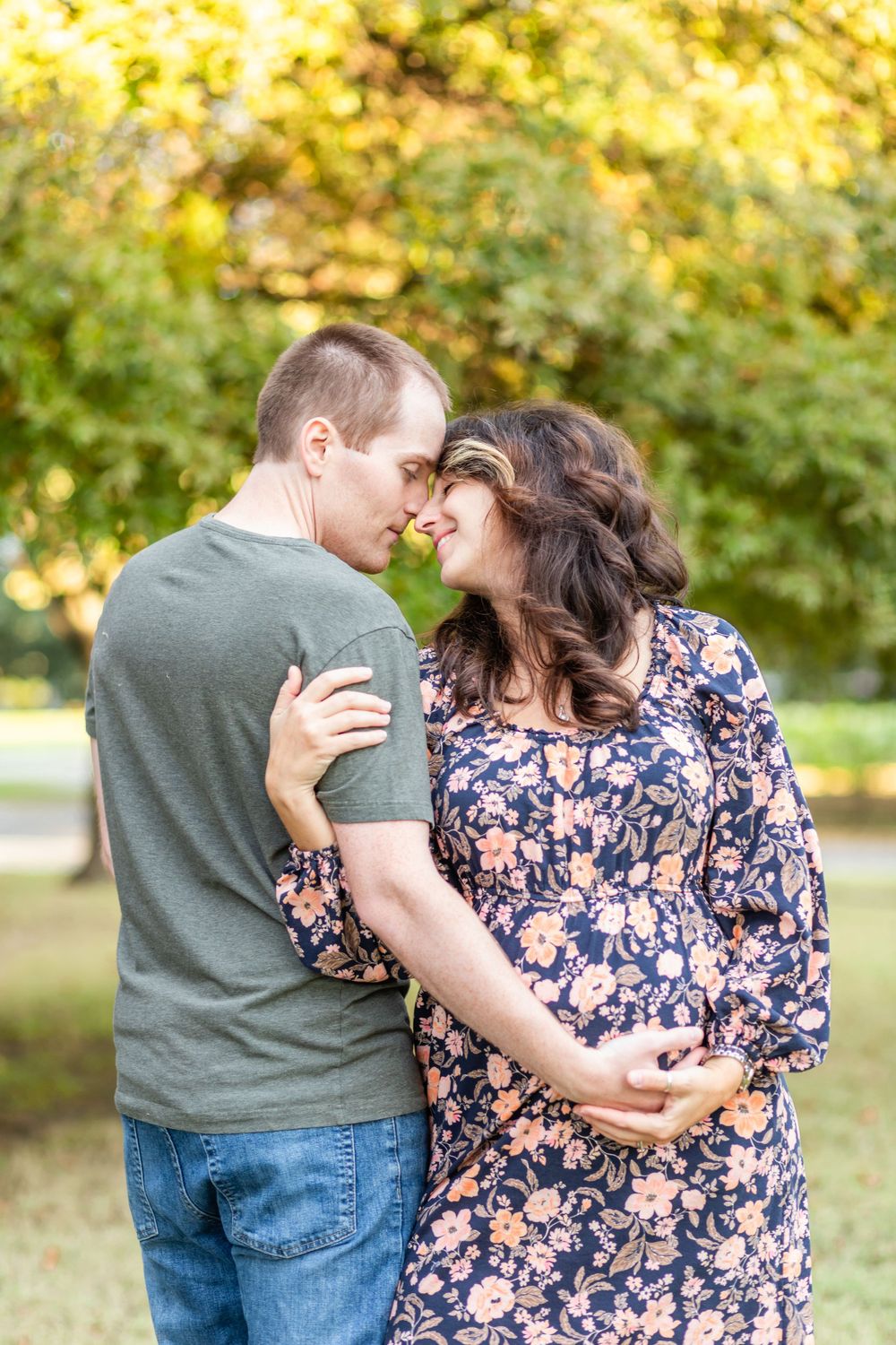 An expectant couple embraces in a park during golden hour.