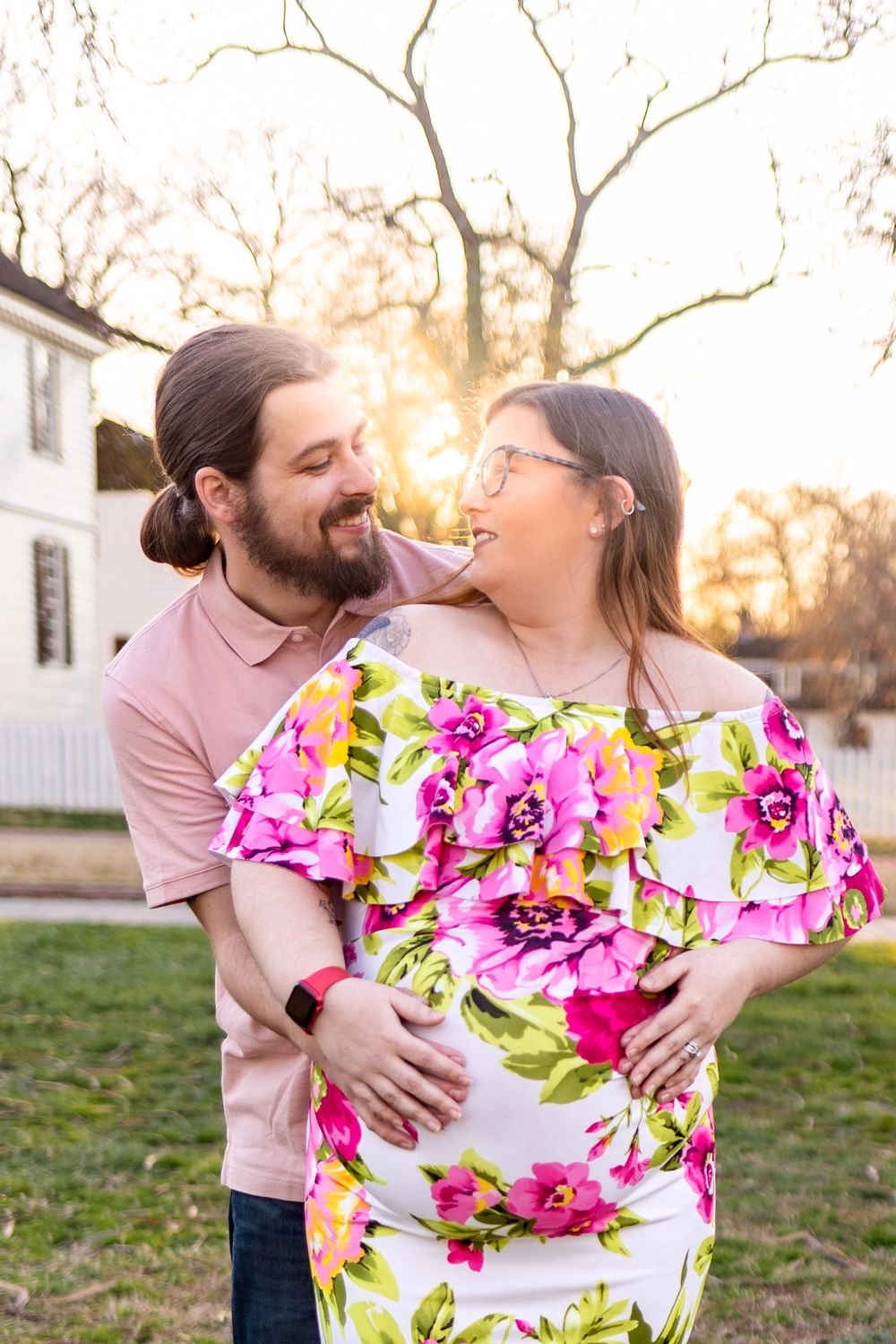 Intimate couple portrait at sunset with pink floral dress against white house background.