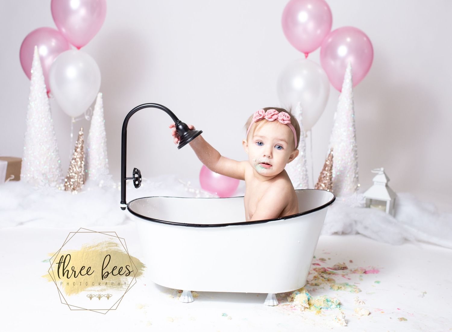 A small child sits in a vintage-style bathtub surrounded by pink and white balloons for a first birthday photoshoot.