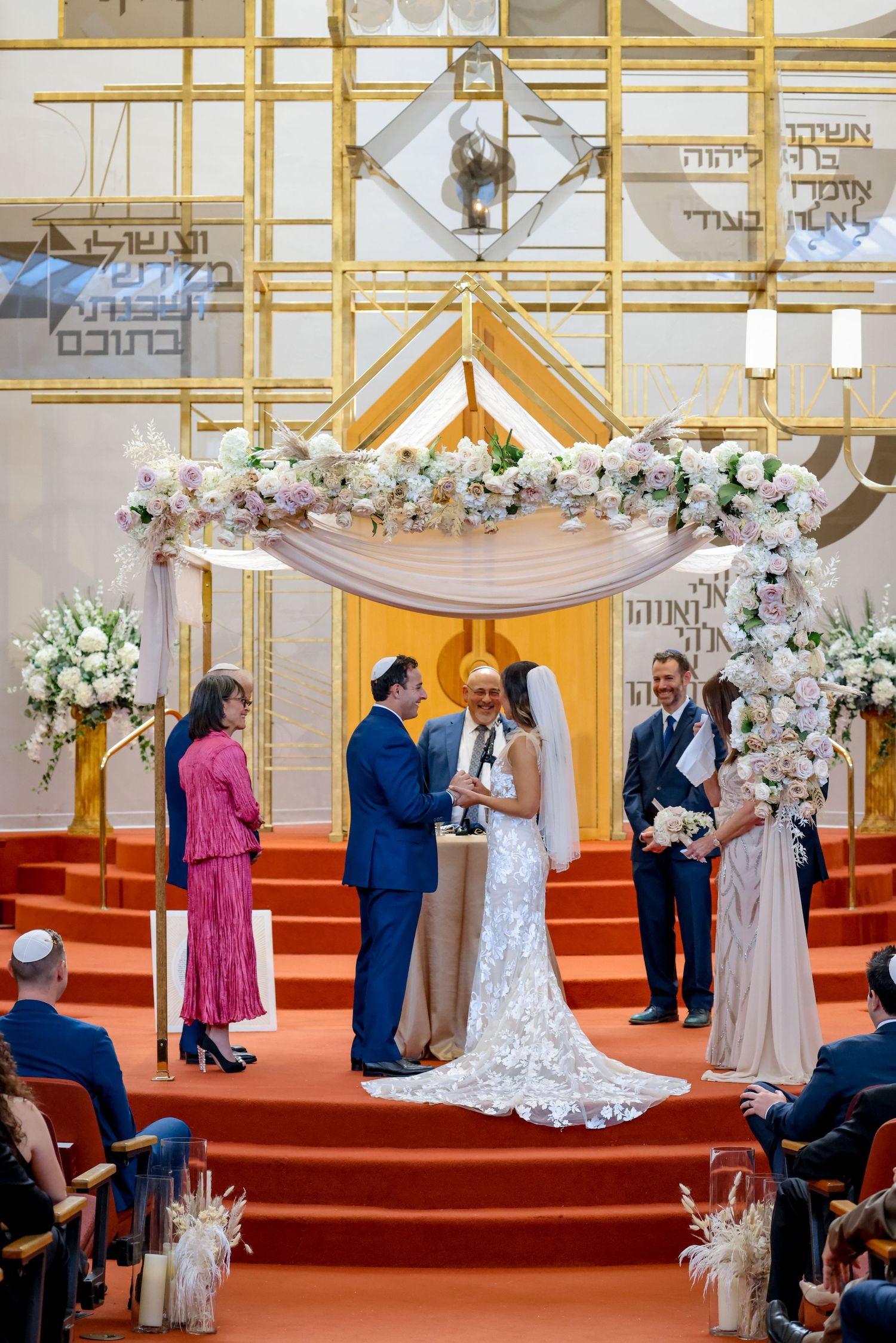 Jewish wedding ceremony under an ornate floral chuppah with gold accents and red carpeting.