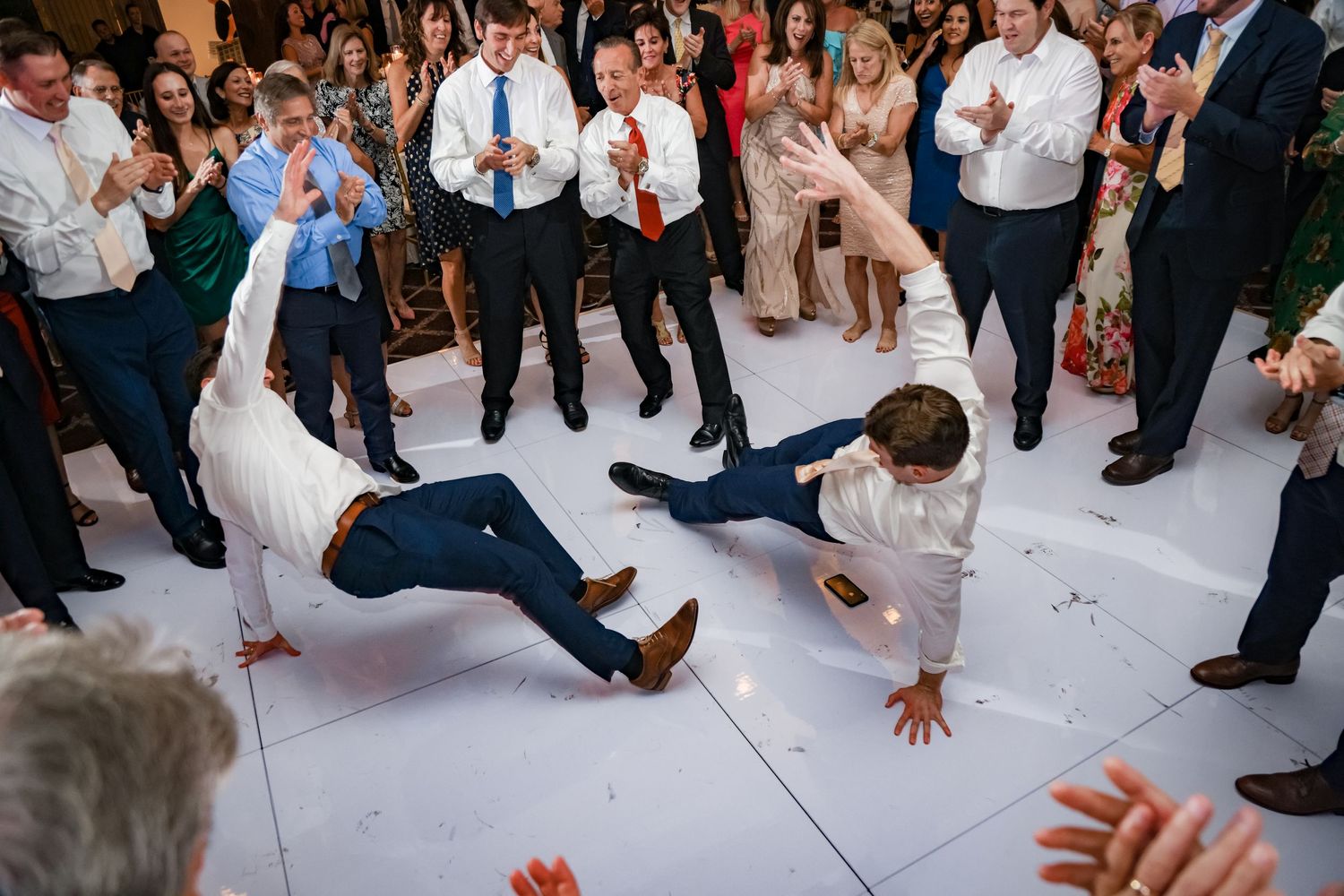 Wedding guests cheer as two people perform breakdancing moves on a white dance floor at a reception celebration.