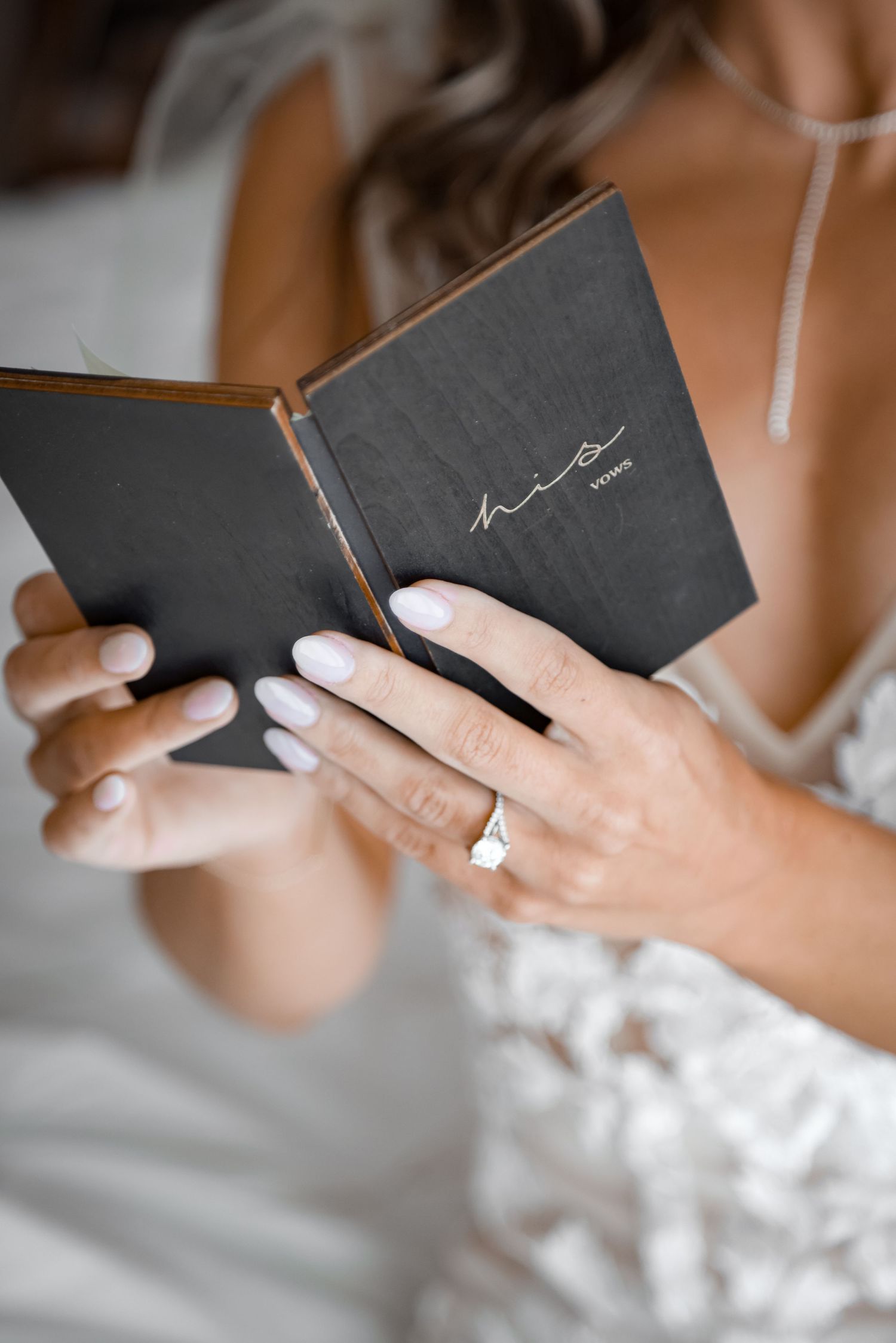 Close-up of hands holding a small black vow book with delicate lace dress details visible.