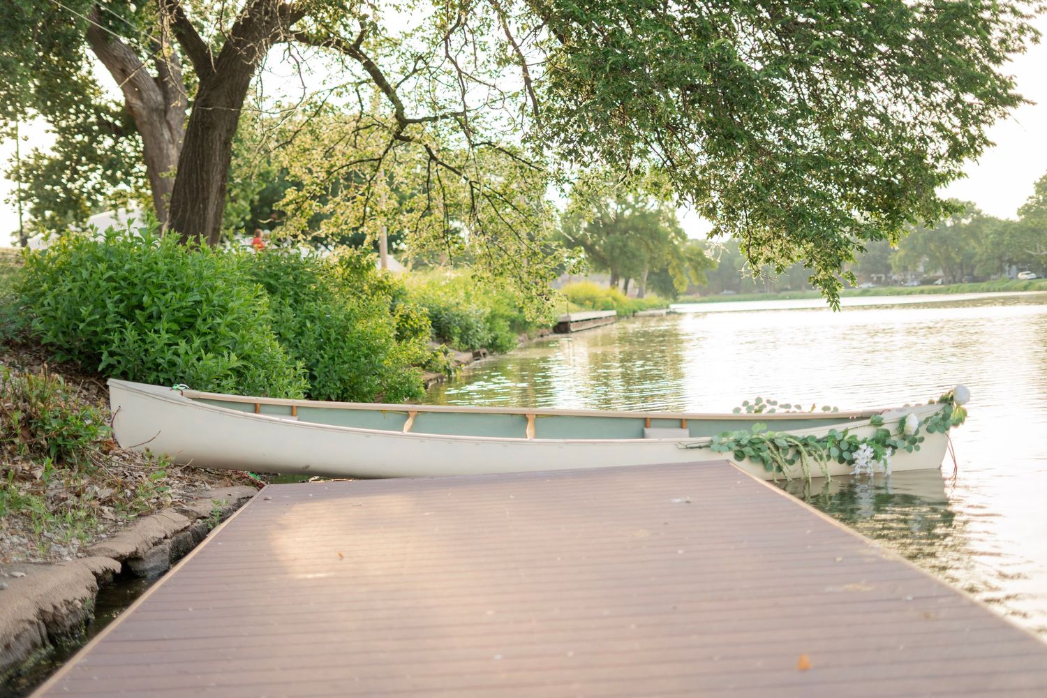 White canoe with floral garland moored at a wooden dock beside a lush green riverbank.