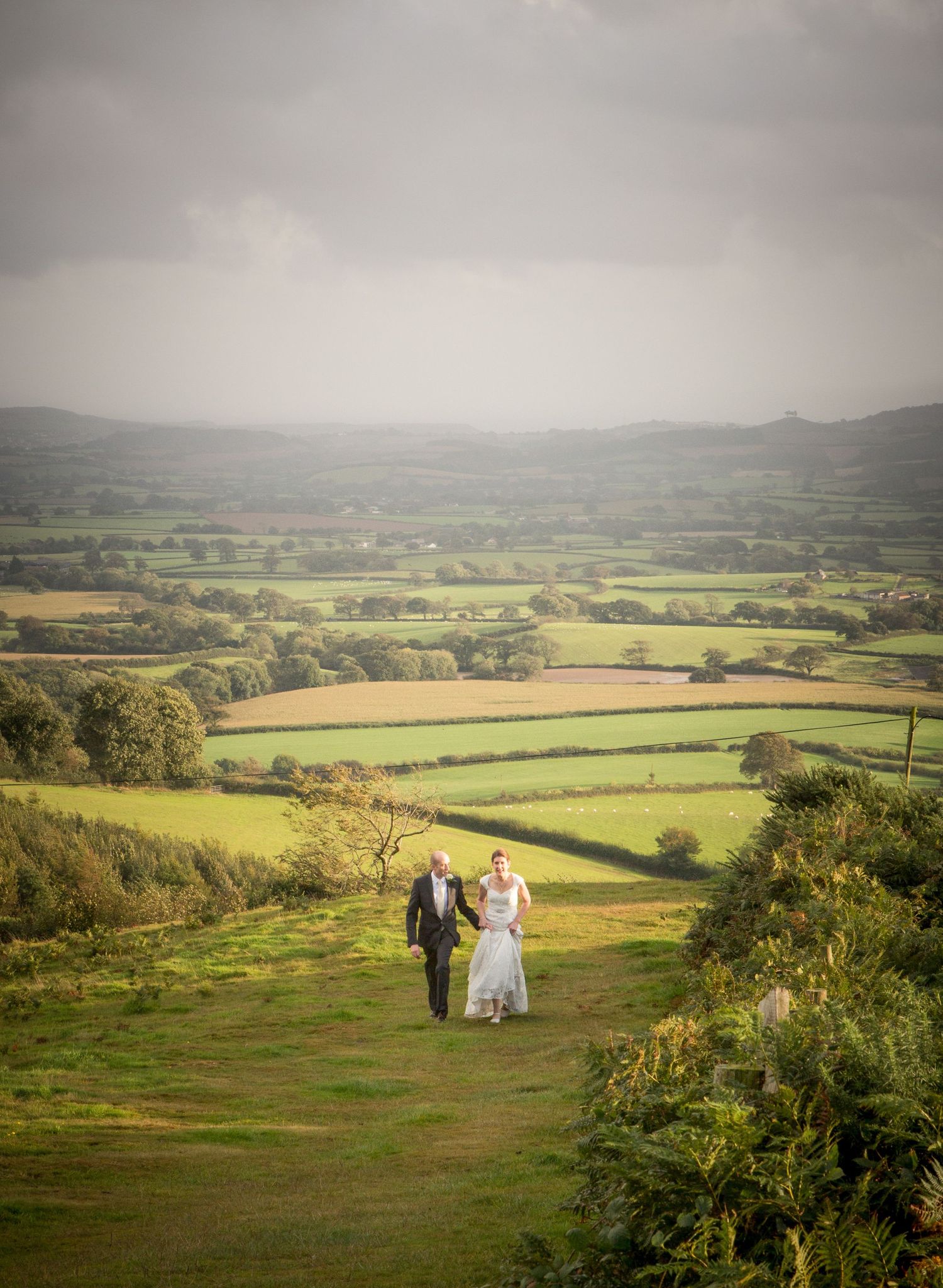 A wedding party stands on a hilltop overlooking rolling green countryside fields.