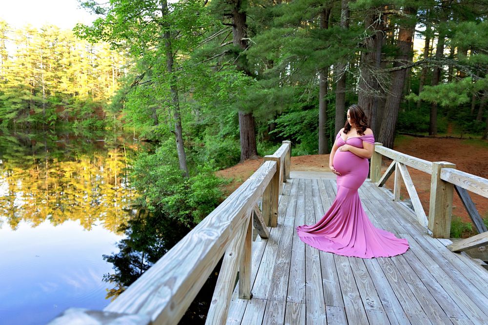 Pregnant woman in pink maternity gown poses on wooden bridge overlooking serene forest lake.