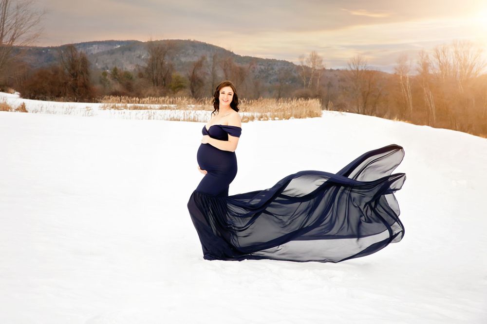 Pregnant woman in navy blue flowing gown poses gracefully in snowy winter landscape.