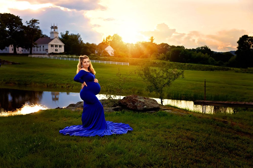 Expectant mother in royal blue maternity gown glows in golden sunset by farm pond.