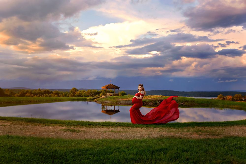 Pregnant woman in flowing red gown poses by reflective pond under dramatic sunset sky.