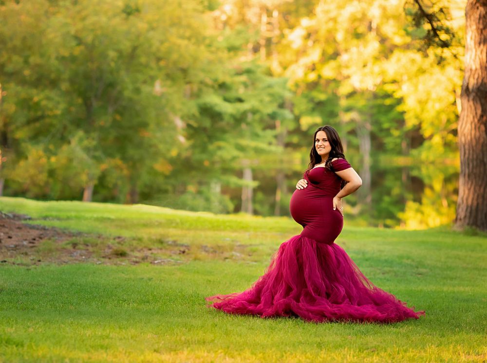 Pregnant woman in burgundy tulle mermaid gown stands on sunlit green park lawn.