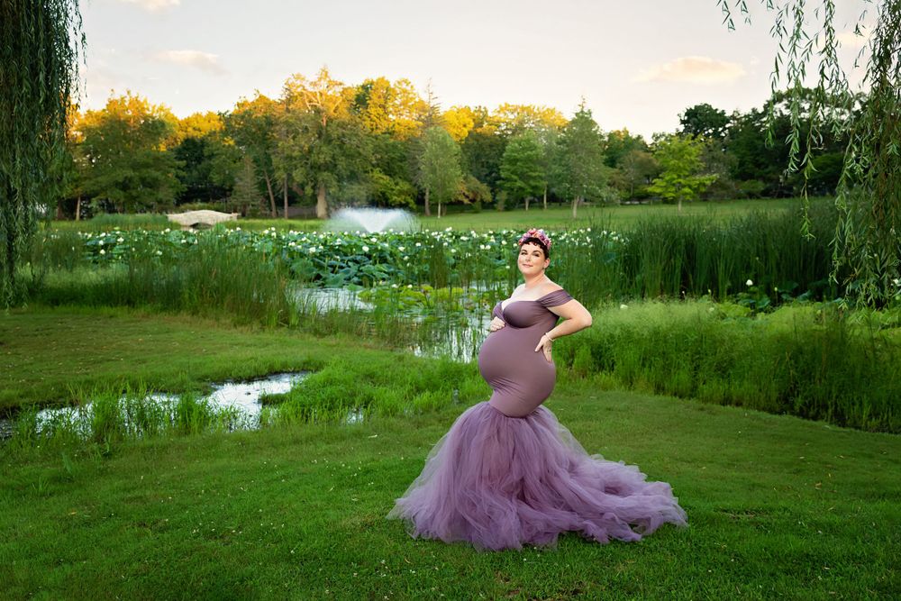 Pregnant woman in purple tulle mermaid gown stands by lily pond in lush green park.