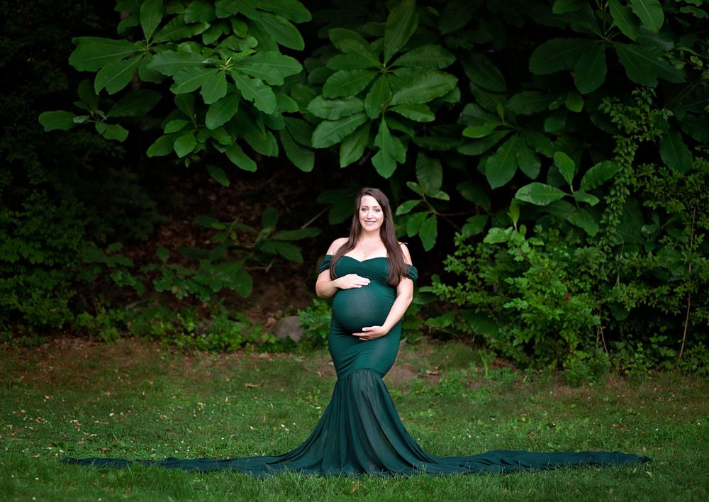 Smiling pregnant woman in dark green mermaid maternity gown poses on lush garden lawn.