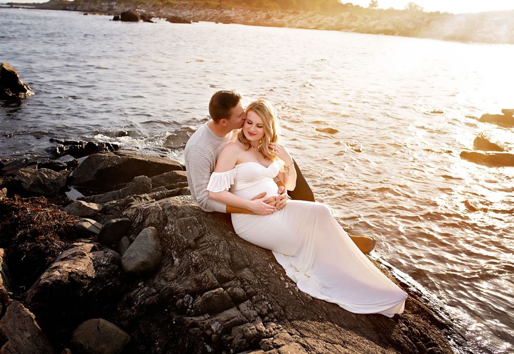 Couple sharing tender moment on rocks by water at sunset, woman in white maternity gown.