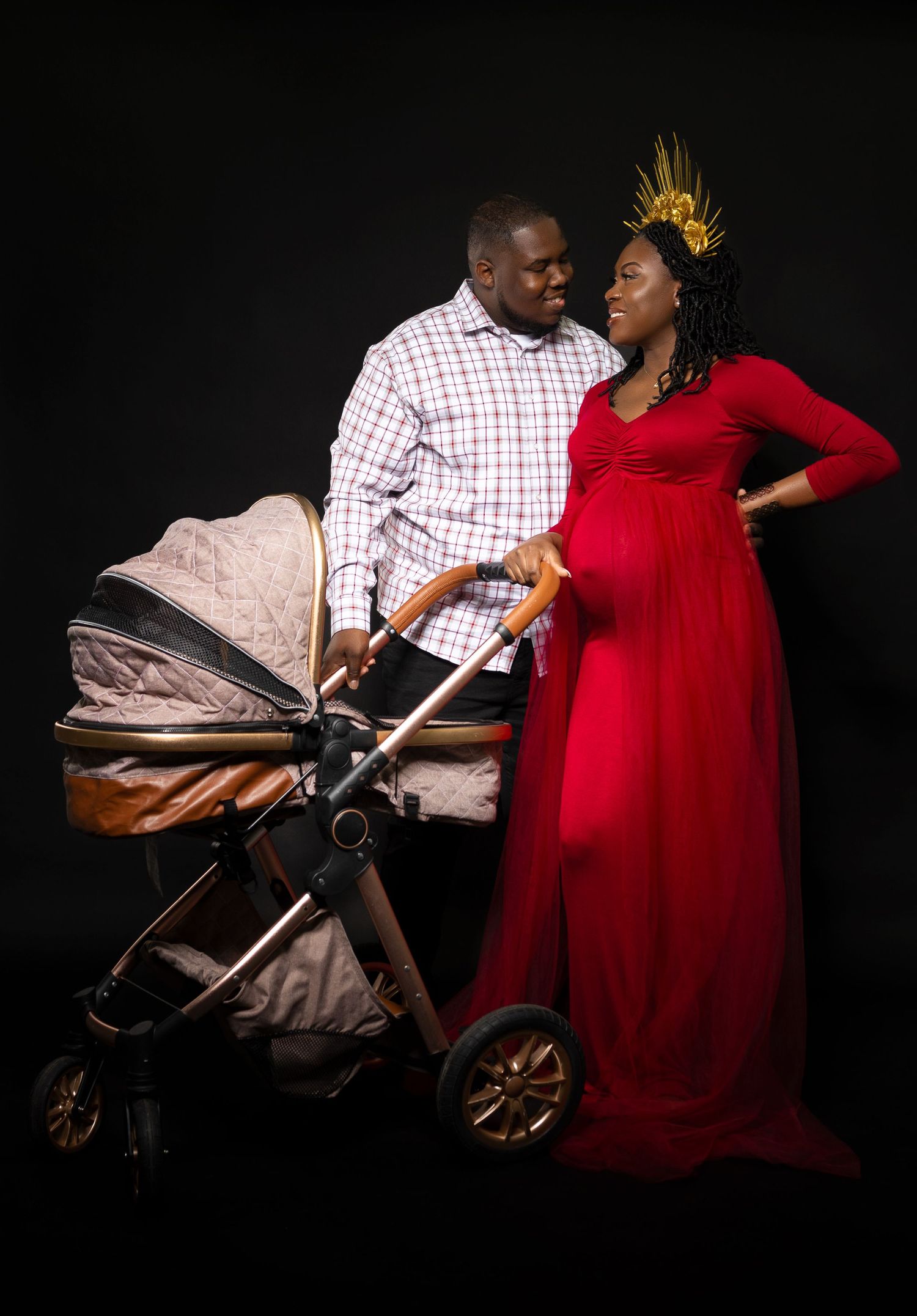 Maternity photo featuring couple in red dress and plaid shirt with baby stroller against black backdrop.