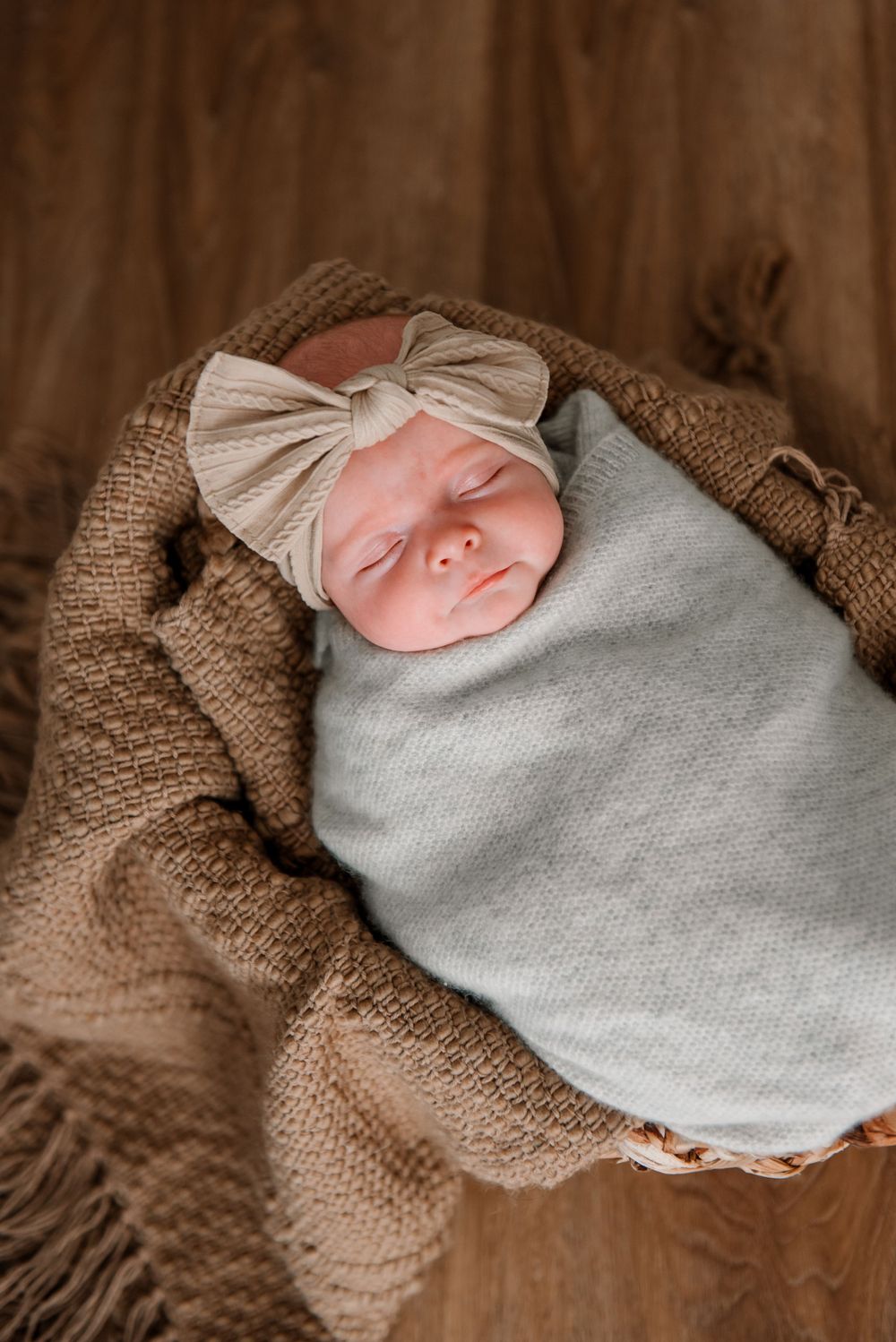 A sleeping infant wrapped in a gray blanket and brown knit wrap on wooden flooring.