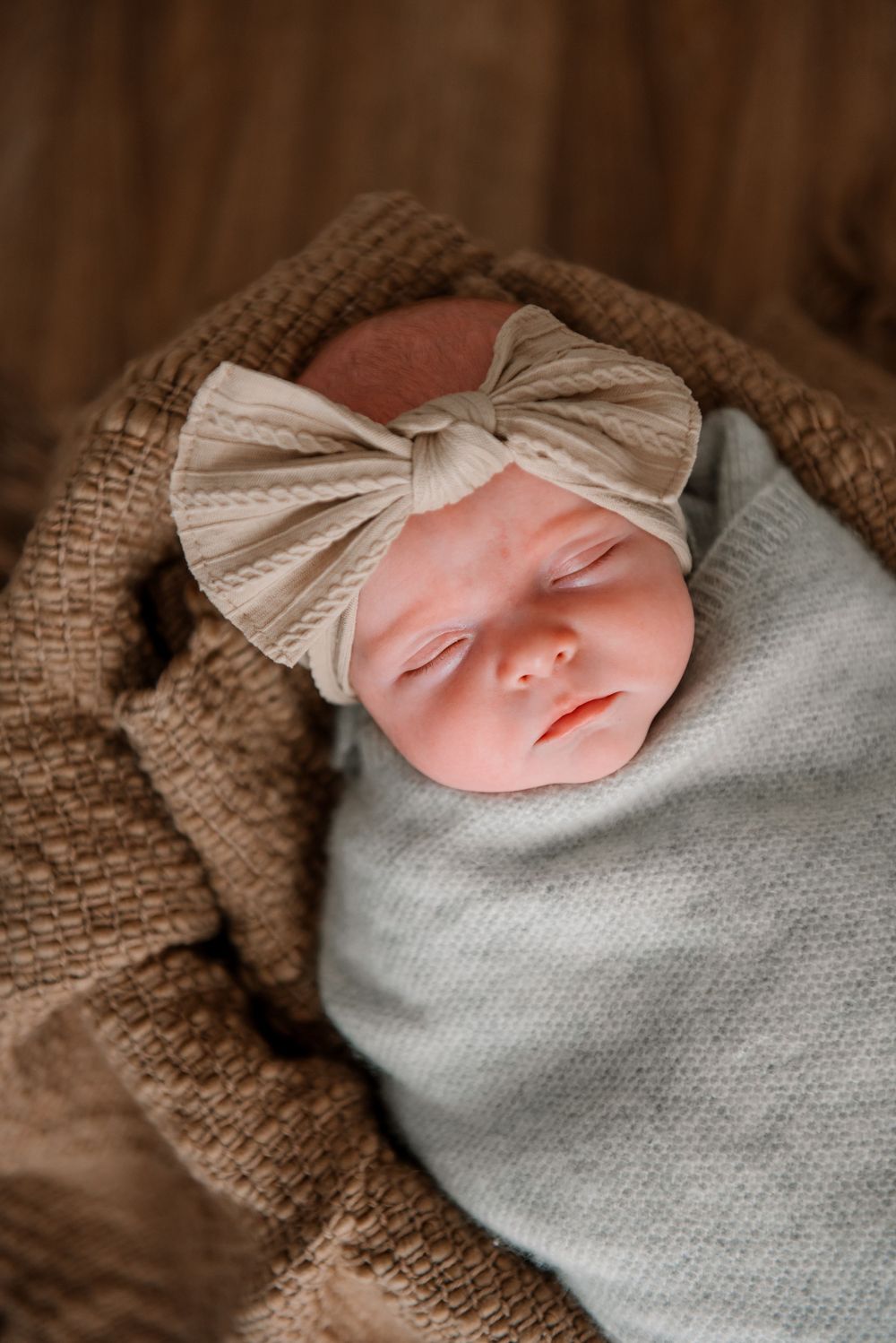 A peaceful sleeping newborn wrapped in gray with a bow headband on brown textured fabric.