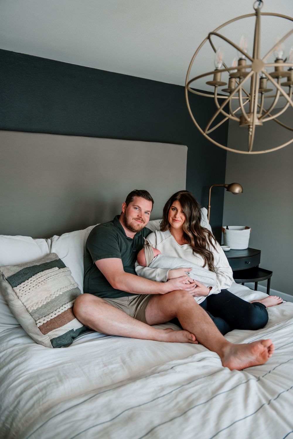 A couple relaxes together on a cozy white bed with decorative pillows and a modern chandelier overhead.