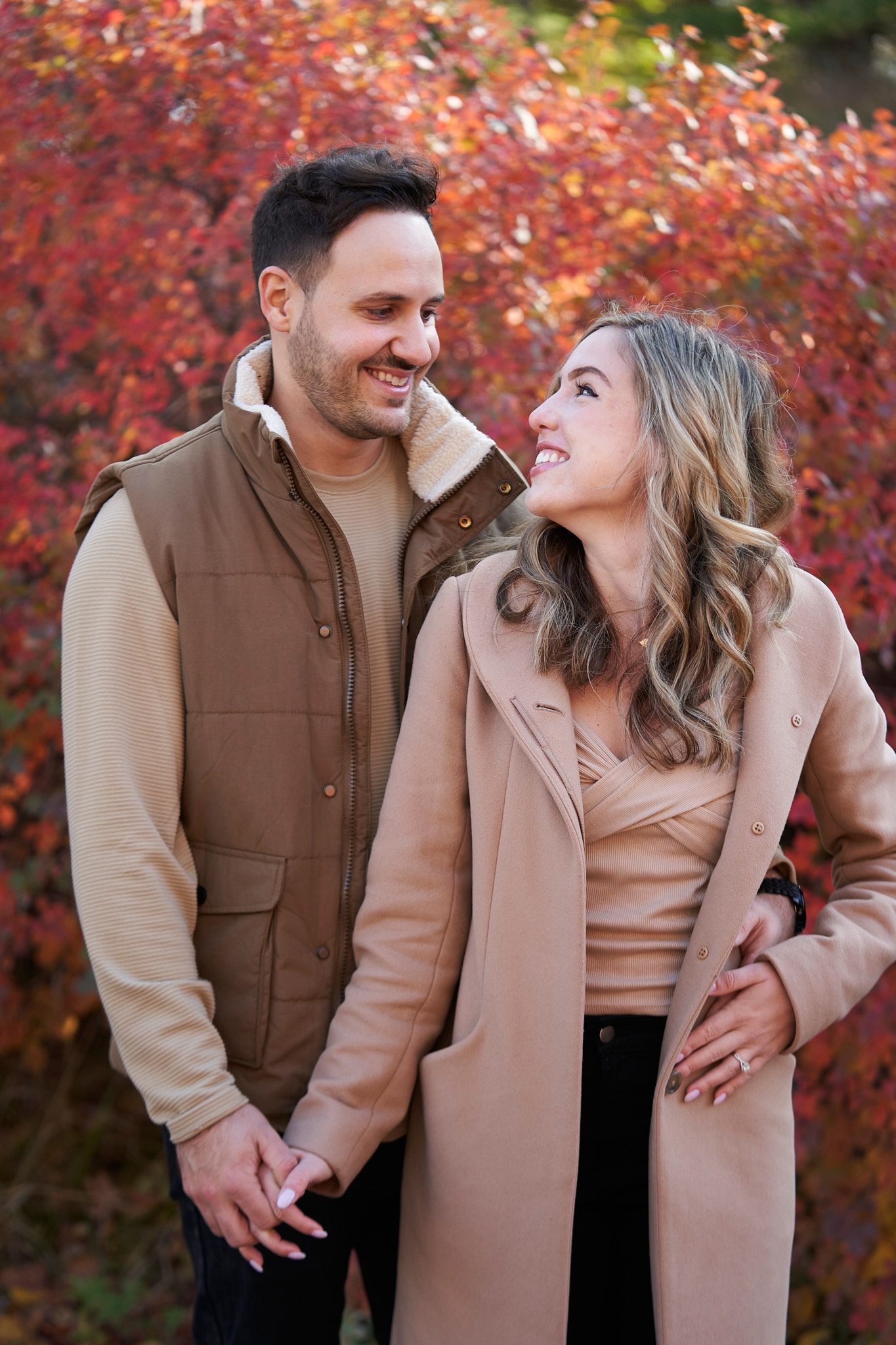 A couple smiles at each other against vibrant red fall foliage wearing coordinating neutral tones.