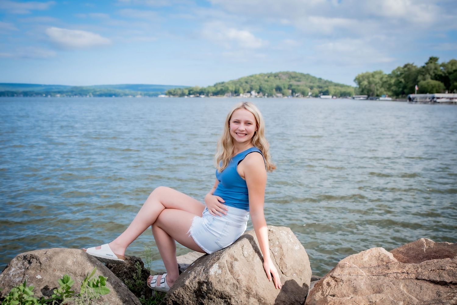 A person poses on large lakeside rocks with blue water and rolling hills in the background.