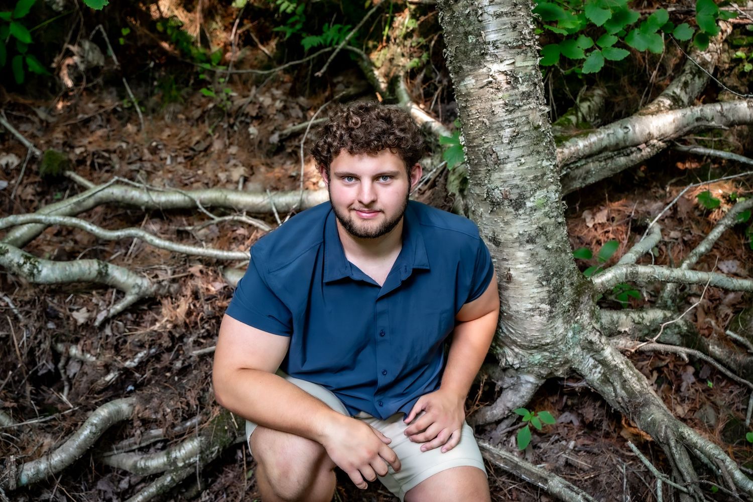A person sits casually against a tree trunk with exposed roots in a natural woodland setting.