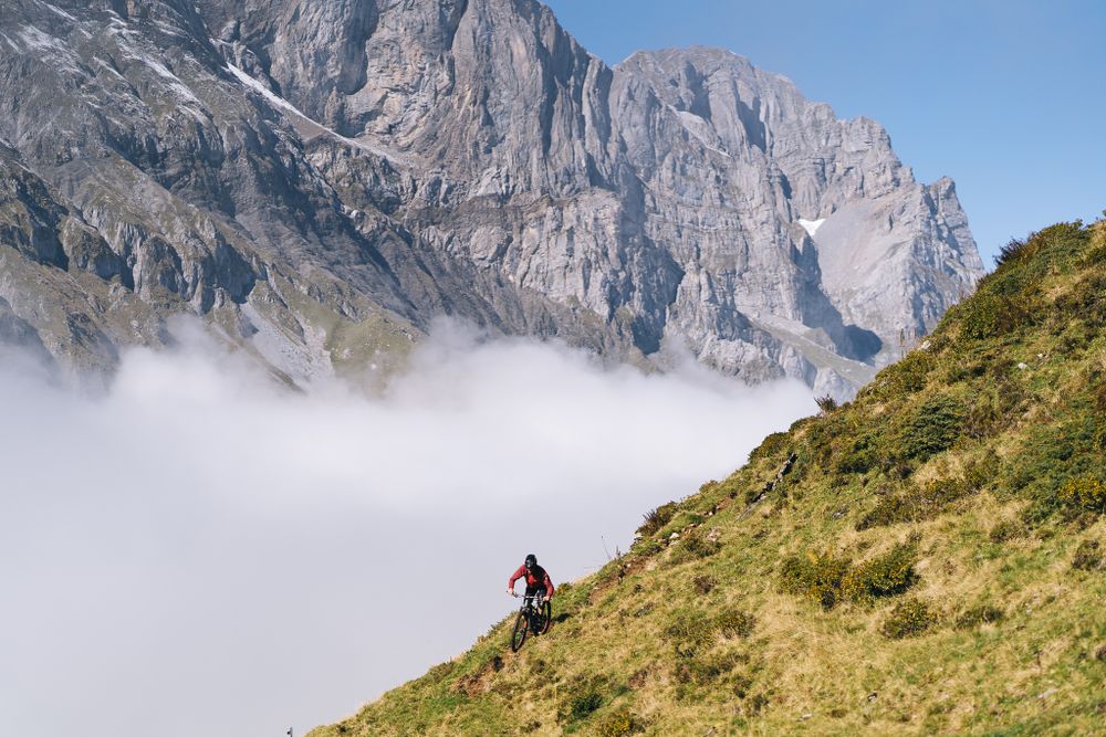 Mountain biker traverses a scenic alpine trail with dramatic rocky cliffs rising through misty clouds.